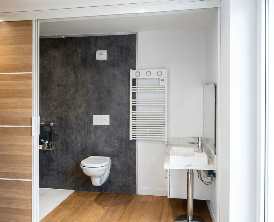 Un &eacute;lectricien professionnel en tenue de travail effectuant des travaux de mise aux normes &eacute;lectrique dans un salon d'une maison en r&eacute;novation. En arri&egrave;re-plan, visible mais l&eacute;g&egrave;rement flou, on distingue d'autres &eacute;l&eacute;ments de chantier sugg&eacute;rant des travaux multi-domaines&nbsp;: des tuyaux de ventilation, des panneaux d'isolation thermique contre un mur, et une partie de porte entrouverte donnant acc&egrave;s &agrave; une salle de bain. La sc&egrave;ne doit transmettre l'id&eacute;e d'un professionnel g&eacute;rant l'ensemble d'un projet de r&eacute;novation globale. Lumi&egrave;re naturelle ou semi-professionnelle, ambiance de chantier r&eacute;el.