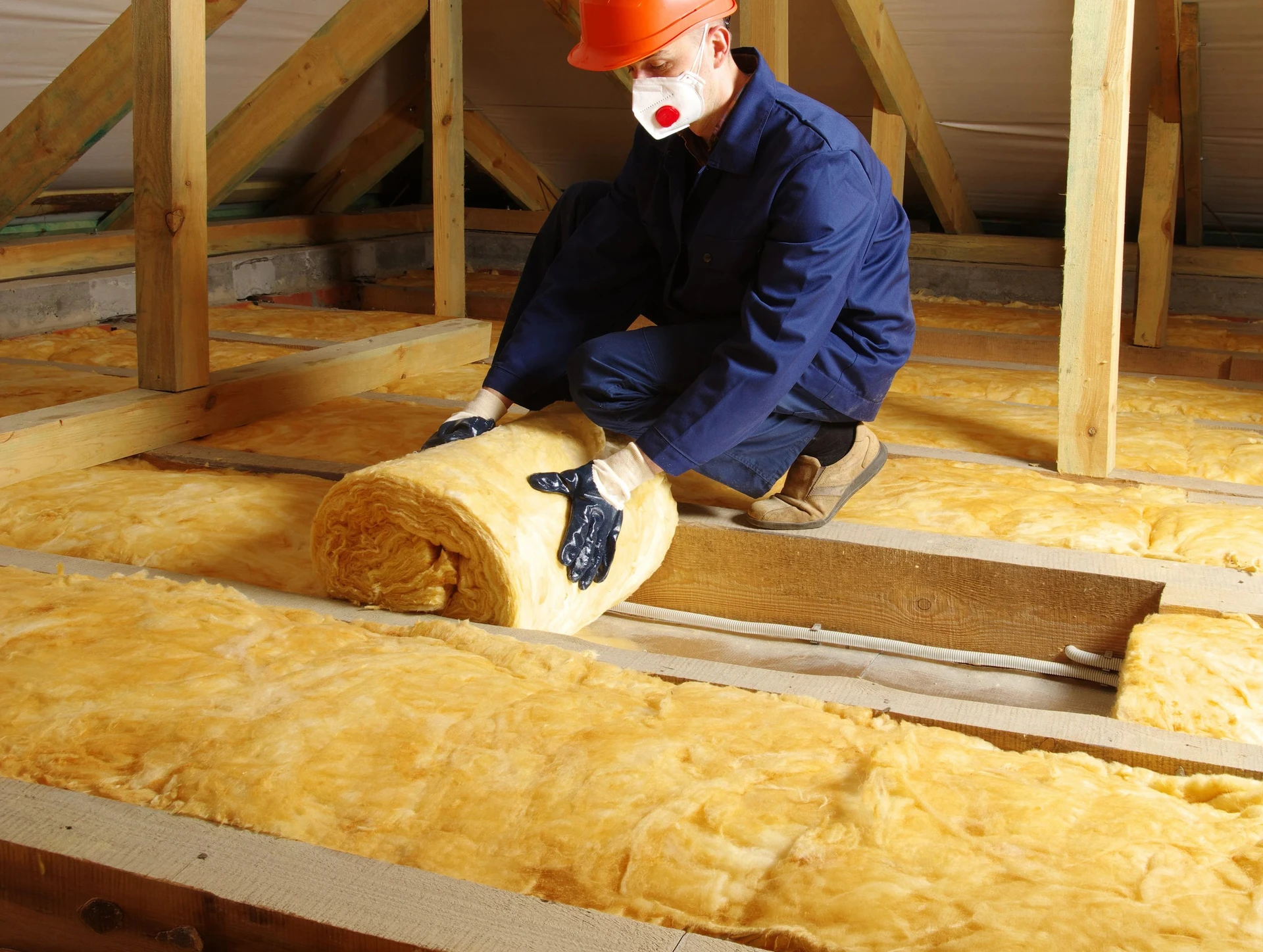 Ouvrier en combinaison bleue et masque respiratoire installant de l'isolation thermique dans les combles d'une habitation